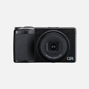 Fotocamera compatta Ricoh GR IV nera vista di fronte con l'obiettivo esteso, la scritta GR LENS f=18.3mm 1:2.8 sulla ghiera e il logo GR sul frontale