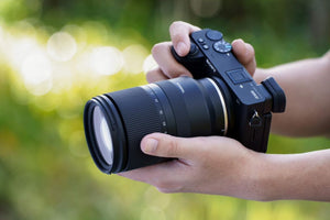 Tamron 17-70mm f/2.8 montato su fotocamera Sony impugnata a due mani, sfondo verde sfocato